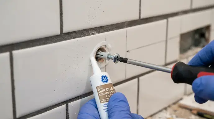 Applying waterproof silicone caulk around a drilled tile hole before driving heavy-duty screws into a wall stud